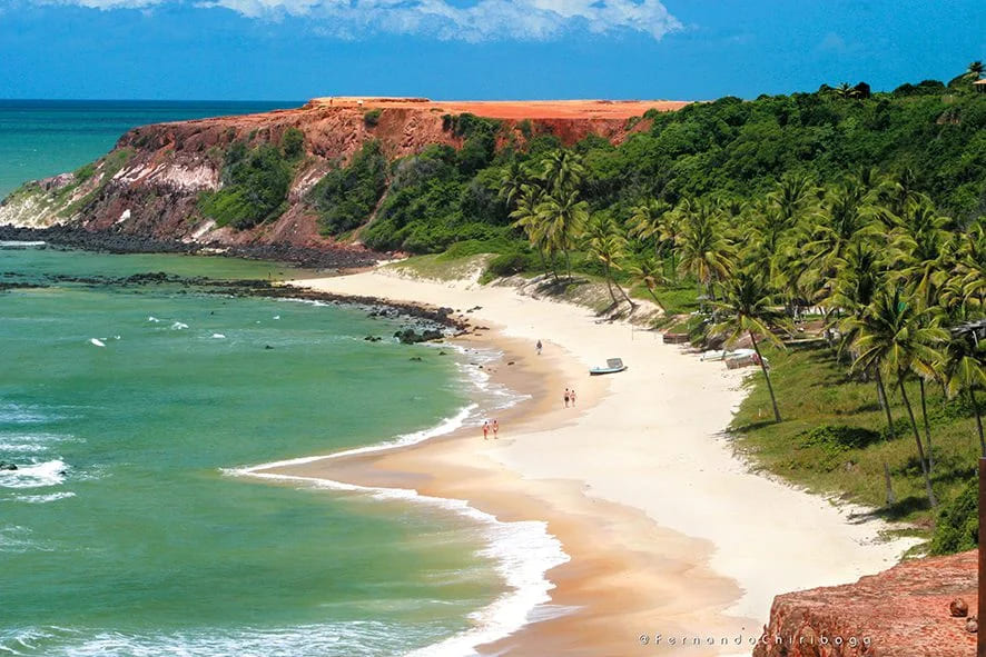 Vista panorâmica da Praia do Amor no Rio Grande do Norte, com suas falésias avermelhadas, coqueiros, areia branca e águas verde-azuladas, destacando o formato de coração da praia.